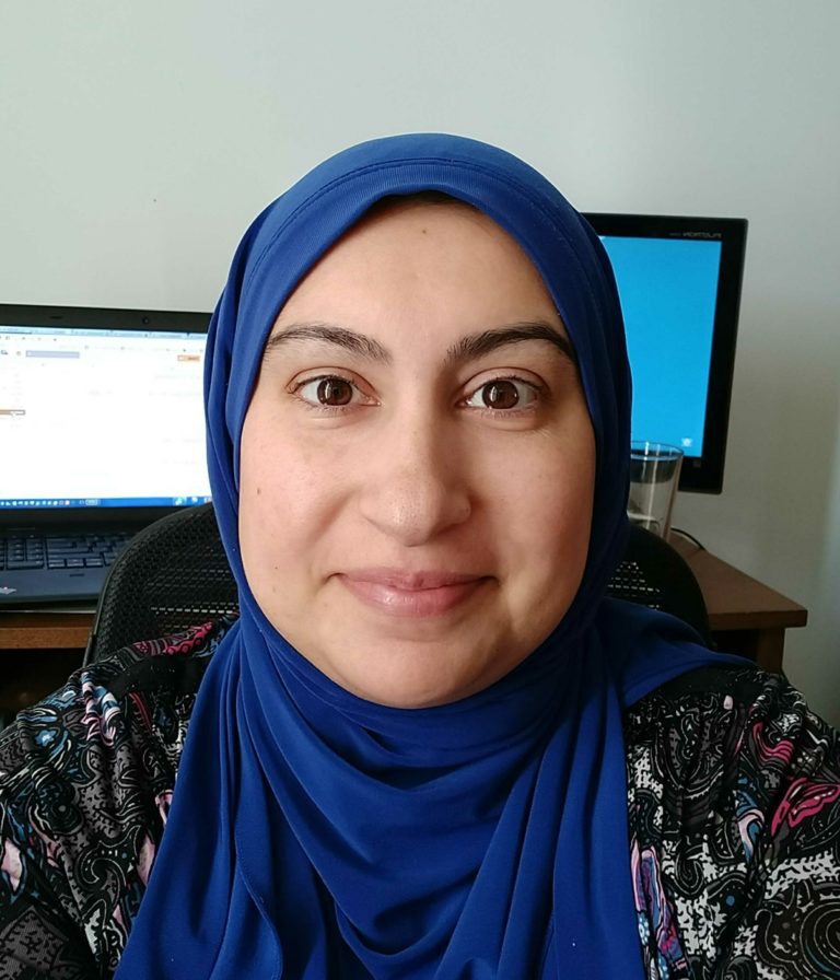 profile picture of muslim woman in tech, Sadiya Zackria. A smiling woman wearing hijab sitting in front of two computer monitors
